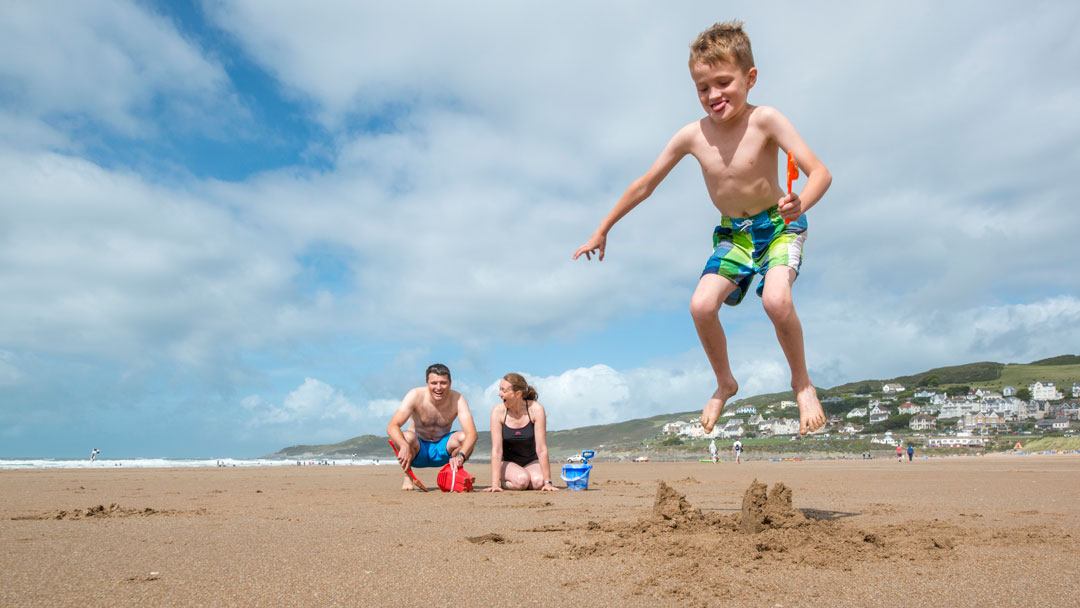 boy jumping on a sandcastle he made whilst holding a spade as his amused parents watch on with Woolacombe hills and Atlantic Ocean in background